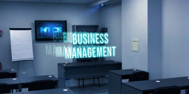 Un'aula moderna con una TV montata a parete che mostra "NTCS" e una fila di banchi con sedie di fronte alla cattedra dell'insegnante. Le parole "BUSINESS MANAGEMENT" sono ben visibili in lettere blu luminose davanti alla cattedra, promuovendo corsi di business e management a Napoli e provincia