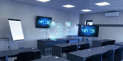 Un'aula moderna con file di banchi e sedie per il corso Progettisti, installatori e manutentori di sistemi elettrici ed elettronici a Napoli. Ci sono due grandi schermi che mostrano un'interfaccia digitale etichettata "NTCS", su misura. Una proiezione olografica al centro mostra varie icone che rappresentano la tecnologia e l'elettronica. La stanza è dotata di lavagna a fogli mobili, finestra e lampada da soffitto.