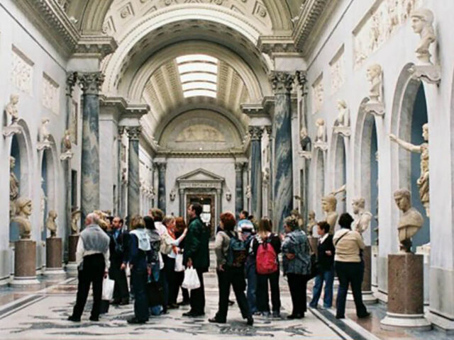 Un gruppo di turisti si trova nel corridoio decorato di un museo, guidato da un Operatore dei Servizi di Custodia ed Accoglienza Museale. I turisti sono circondati da sculture classiche. Il soffitto presenta disegni intricati e grandi lucernai che lasciano entrare la luce naturale esaltando la grandiosità del pavimento in marmo .