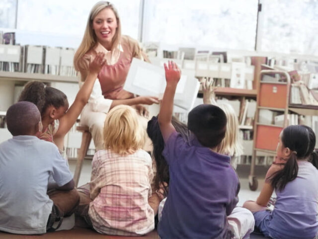 Un'insegnante ( operatore dell'infanzia ), è seduta su una sedia e sorride mentre tiene in mano un libro aperto, interagendo con un gruppo di bambini piccoli seduti sul pavimento di fronte a lei. I bambini hanno le mani alzate e partecipano con entusiasmo. Sullo sfondo si possono vedere scaffali di libri.